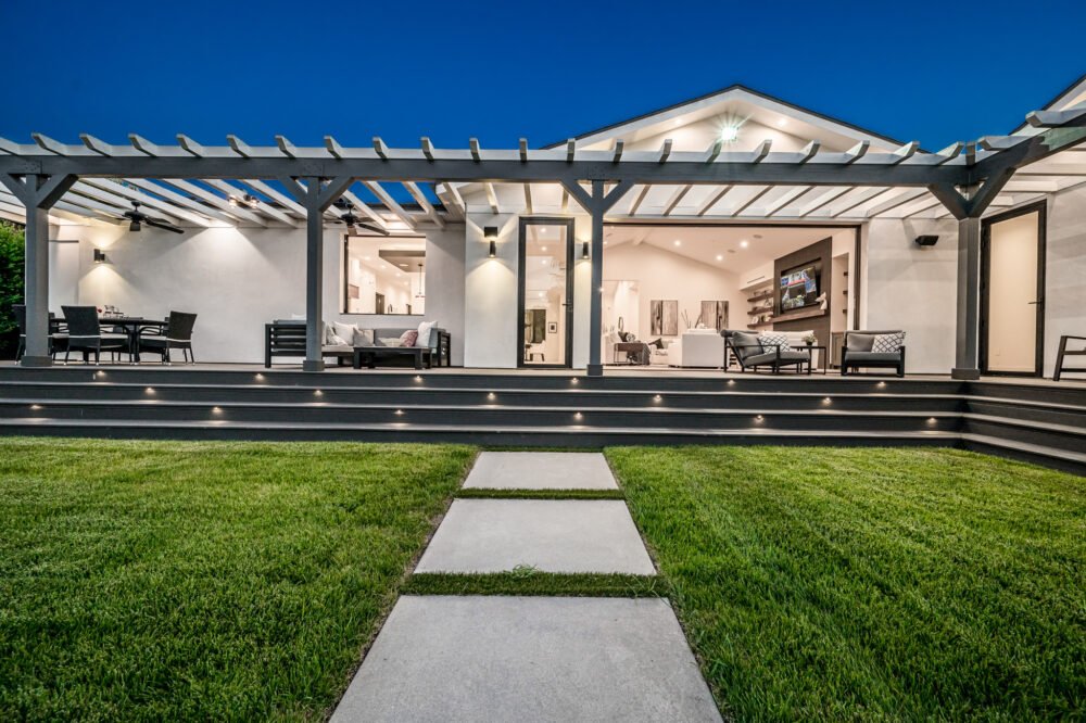 A covered outdoor living space with a wood-plank ceiling, recessed lighting, and a modern seating area overlooking a backyard.